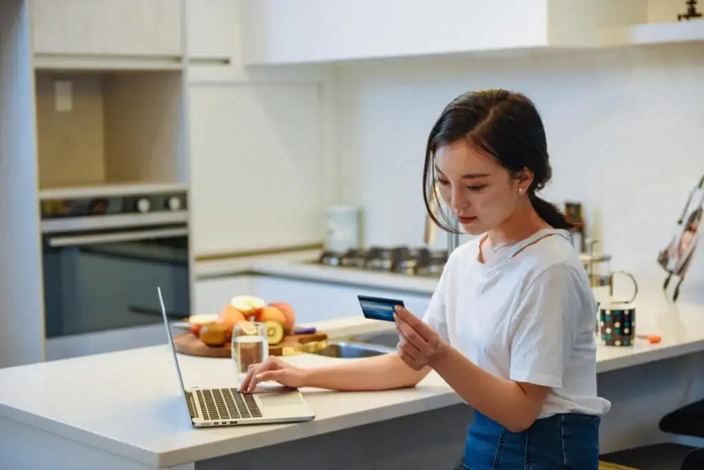 A young woman sits in a modern kitchen using a laptop while holding a credit card, appearing to shop or make an online payment. A bowl of fruit and other kitchen items are visible on the counter beside her.