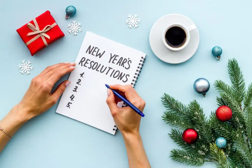 A person writes “New Year’s Resolutions” at the top of a notepad. The desk has a cup of coffee, a red gift box, pine branches with ornaments, and scattered snowflakes, creating a festive atmosphere.