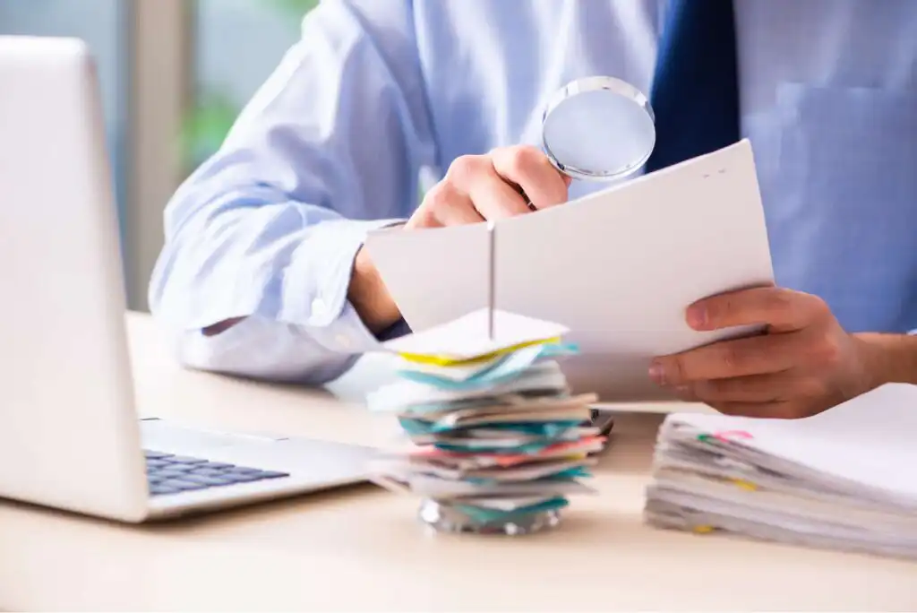 A person in business attire examines documents with a magnifying glass at a desk, with stacks of papers and a laptop nearby.