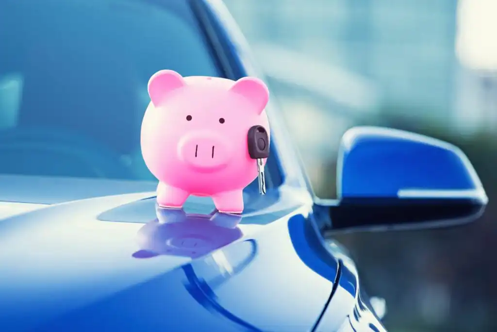 A pink piggy bank with a car key attached stands on the hood of a shiny blue car, symbolizing car savings or financing.