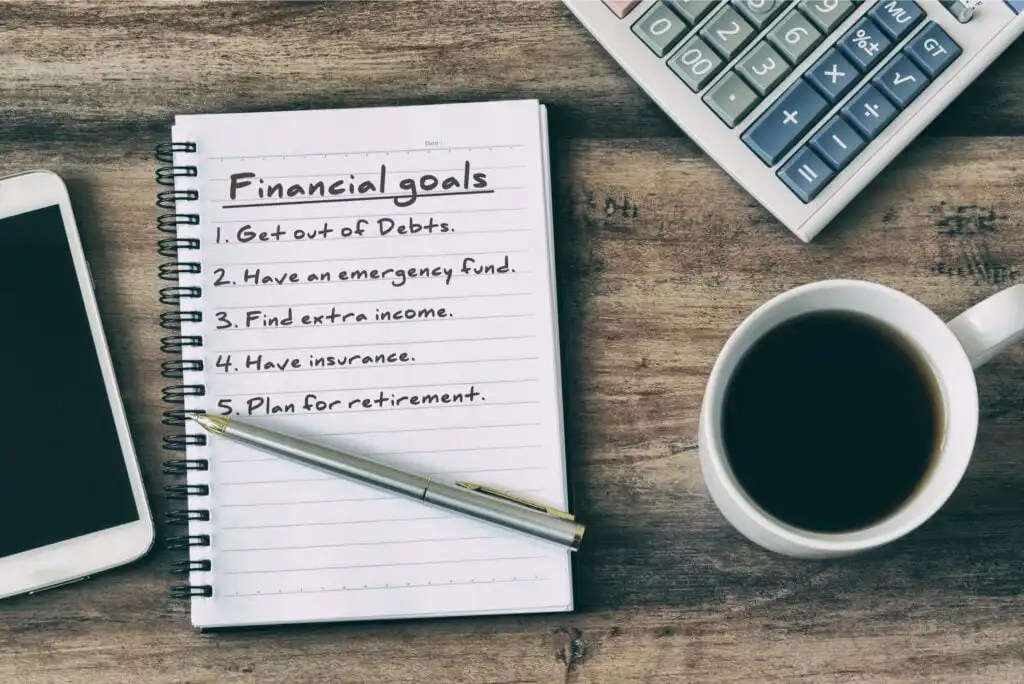 A notebook page titled Financial goals lists five items. Nearby are a pen, a cup of coffee, a smartphone, and a calculator on a wooden surface.