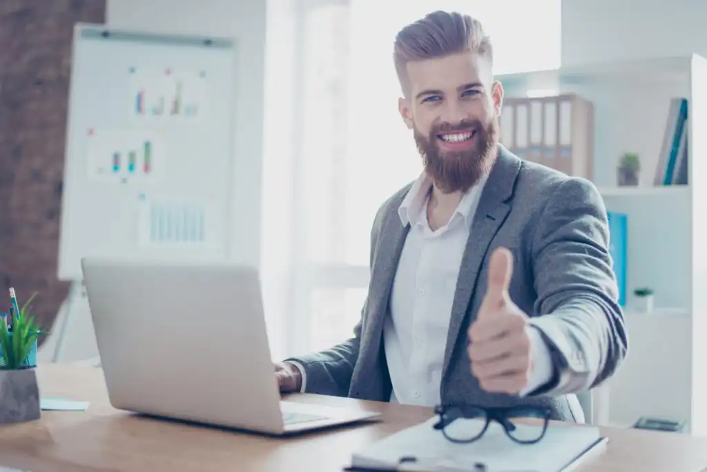 A bearded man in a suit sits at a desk with a laptop, smiling and giving a thumbs up. There are eyeglasses, papers, and a pen on the desk, and charts are visible in the background.