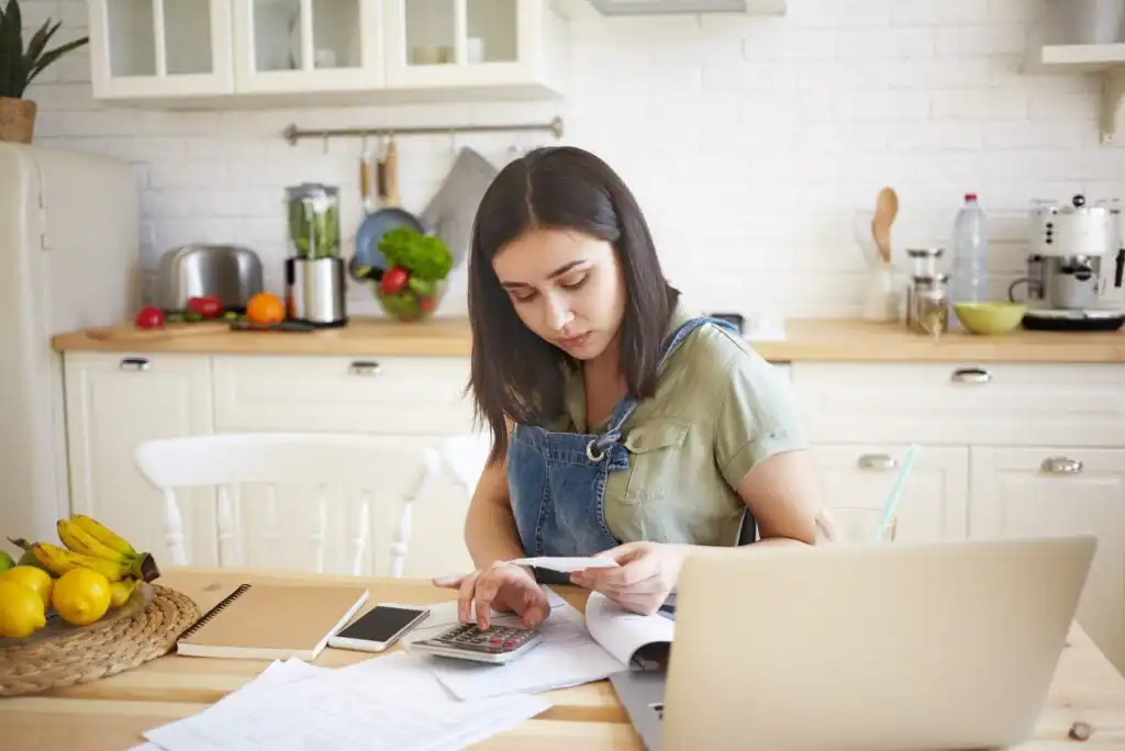 A woman sits at a kitchen table with a laptop, calculator, and papers, appearing focused as she reviews receipts. The kitchen counter behind her holds fresh produce and kitchen appliances.