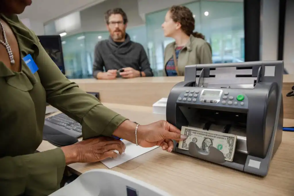 A bank teller feeds a one-dollar bill into a money counting machine at a counter, while two people stand in the background, waiting and talking.