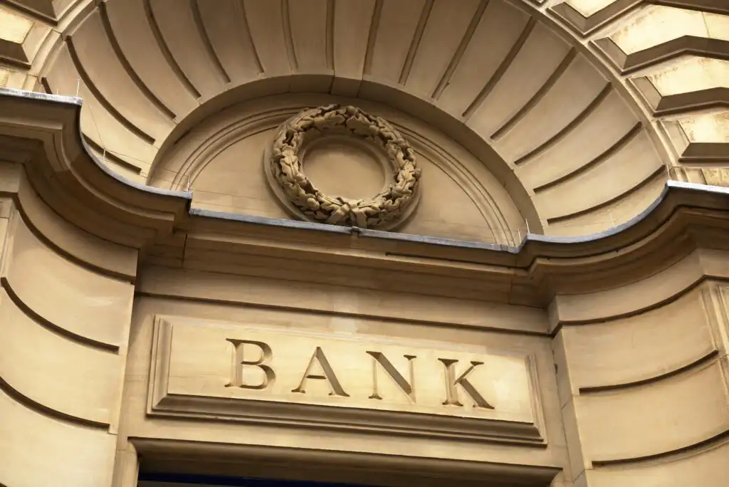 Stone facade of a building with the word BANK engraved above the entrance, an ornate arch, and a carved wreath decoration above the sign.