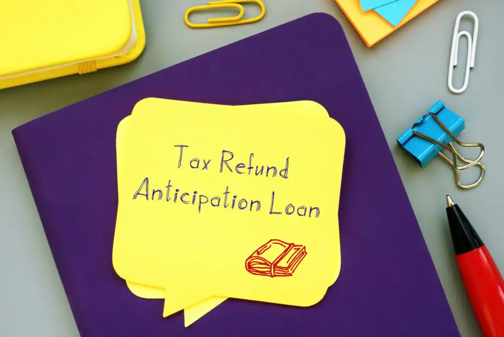 A yellow sticky note on a purple notebook reads “Tax Refund Anticipation Loan” with a small book sketch. Surrounding are paper clips, a pen, binder clips, and other notepads.
