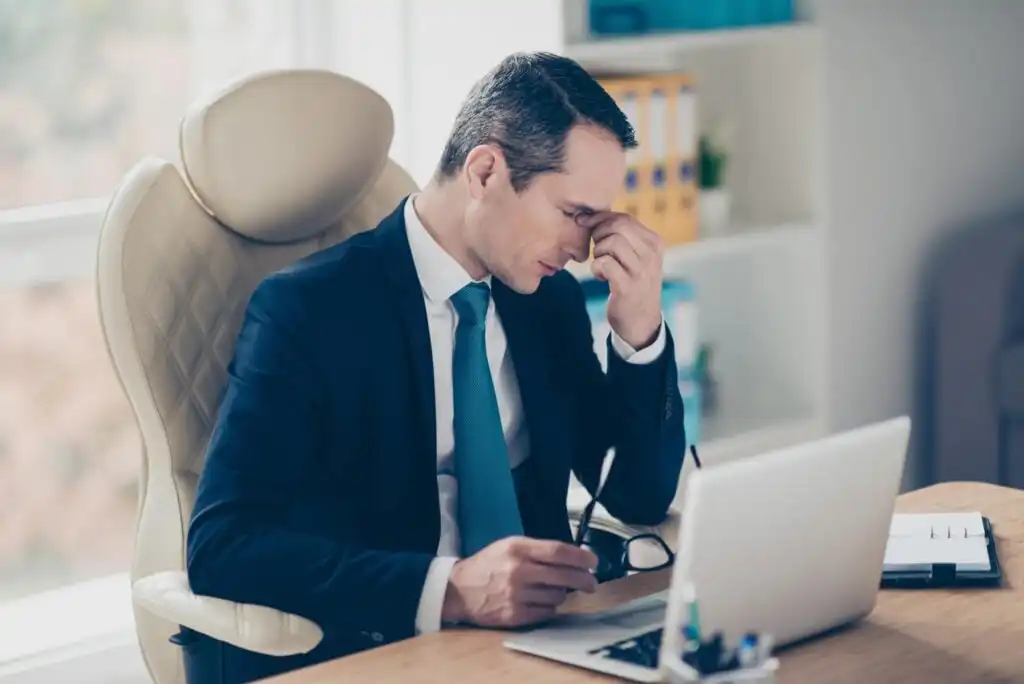 A man in a suit sits at a desk with a laptop, holding his glasses and pinching the bridge of his nose, appearing stressed or tired in an office setting.