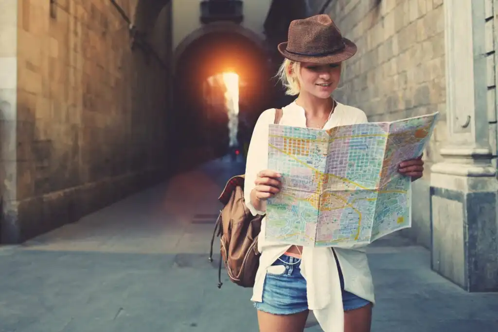 A young woman wearing a hat, backpack, white shirt, and denim shorts stands in a sunlit alley, smiling while reading a map. The street is lined with stone buildings and an archway is visible in the background.
