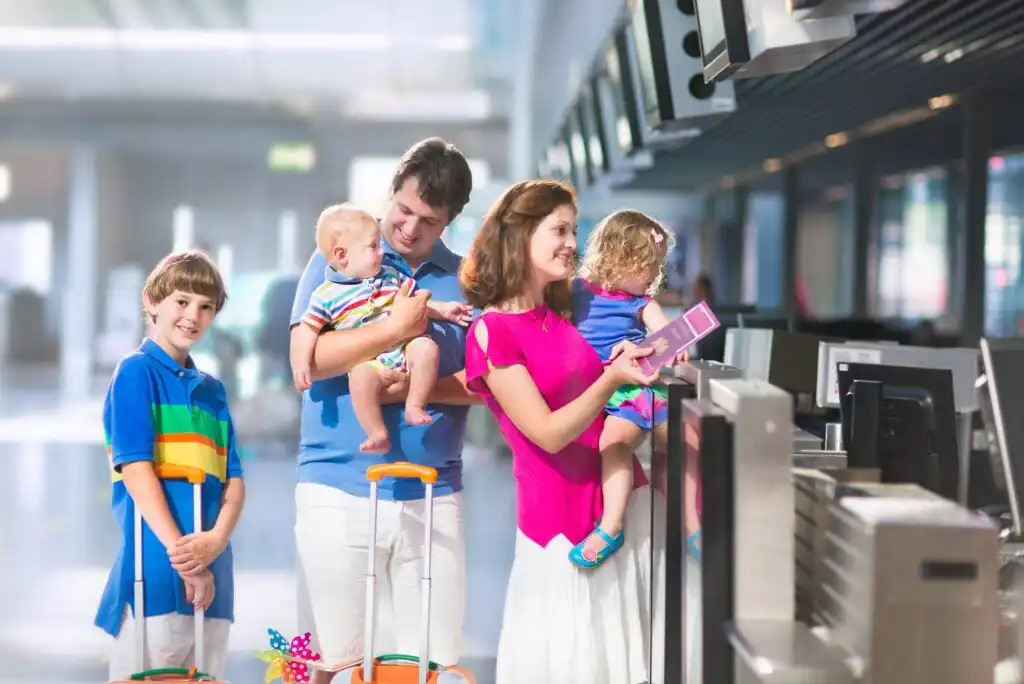 A family of five with luggage stands at an airport check-in counter. The mother holds passports, the father carries a baby, a young girl stands nearby, and an older boy holds a suitcase.