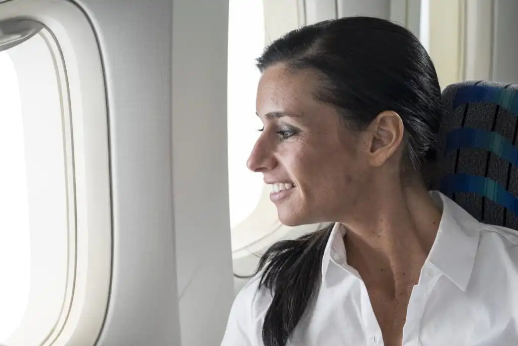 A woman with dark hair in a white shirt sits in an airplane seat, smiling as she looks out the window at bright daylight.