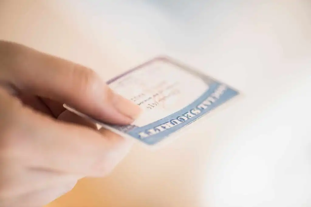 A close-up of a hand holding a Social Security card, with the card slightly out of focus and the background blurred.