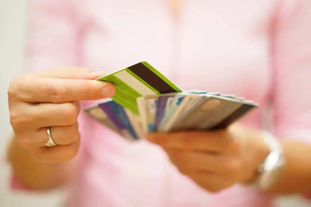 A person wearing a pink shirt holds a fan of credit and debit cards in one hand, selecting one with the other hand. The image is focused on the hands and cards, with the background blurred.