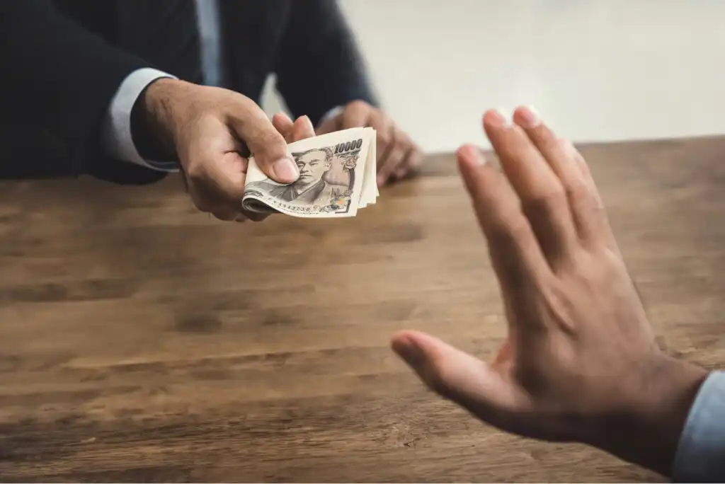 A person in a suit extends a stack of Japanese yen banknotes across a wooden table, while another person raises their hand to refuse the money.