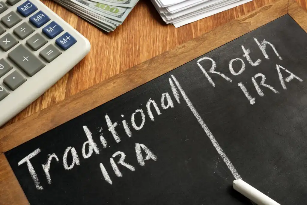 A chalkboard displays Traditional IRA on the left and Roth IRA on the right, divided by a vertical line. Nearby are a calculator, a stack of cash, and papers on a wooden desk.