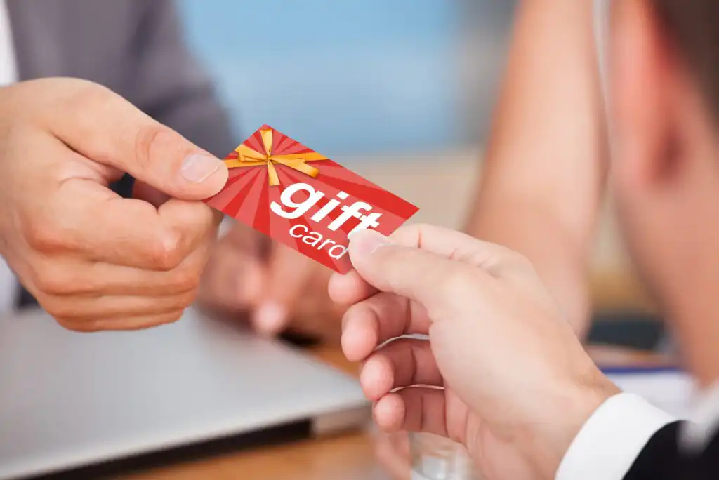 Close-up of two people exchanging a red gift card with a yellow ribbon design. One person is handing the card to another over a table with a laptop in the background.