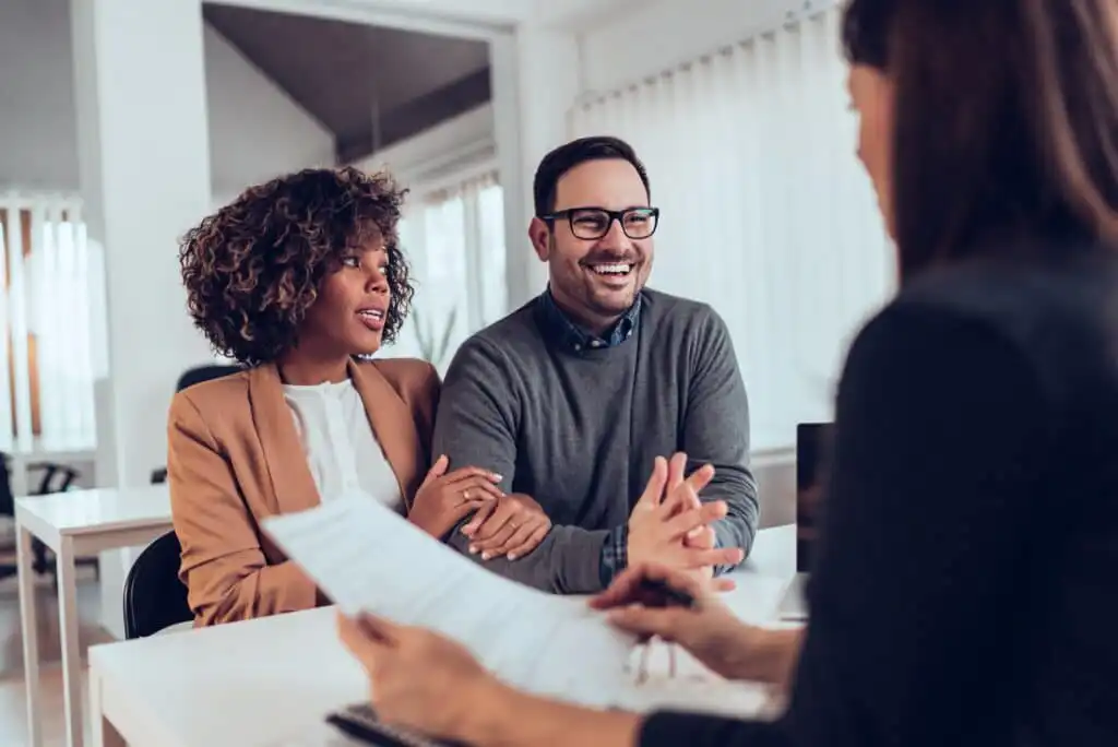 A smiling couple sits at a desk across from a professional holding documents, engaged in conversation in a bright, modern office setting.