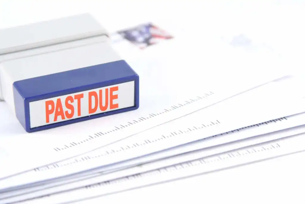 A rubber stamp labeled PAST DUE rests on top of a stack of envelopes and papers, indicating overdue bills or payments. The background is white and the focus is on the stamp and documents.