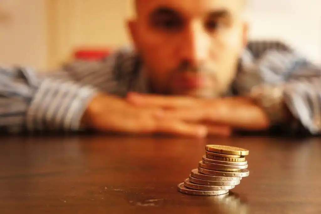 A man rests his chin on his folded hands and looks thoughtfully at a small stack of assorted coins on a wooden table in front of him, with the coins in focus and the man blurred in the background.