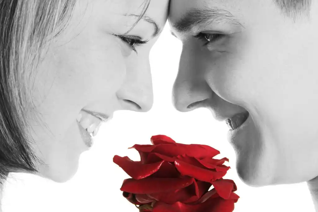 A close-up of a smiling couple touching foreheads, holding a single red rose between them. The image is mostly black and white, except for the vibrant red rose.