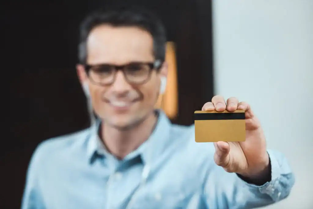A smiling man wearing glasses and a light blue shirt holds a gold credit card toward the camera. The card’s magnetic stripe is visible, and the background is out of focus.