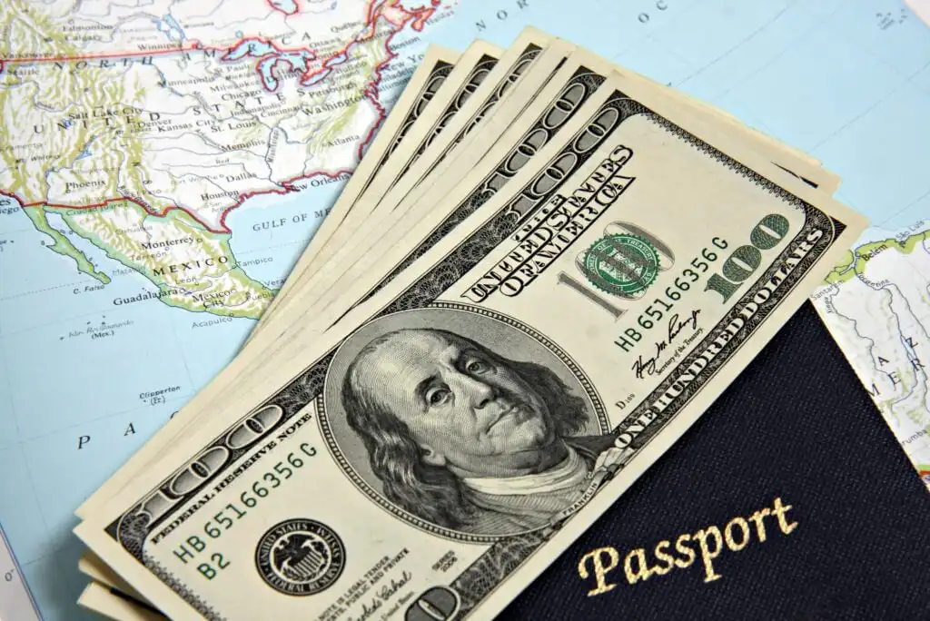 Five $100 bills spread out next to a dark blue passport on top of a colorful map of North America, suggesting themes of travel and finance.