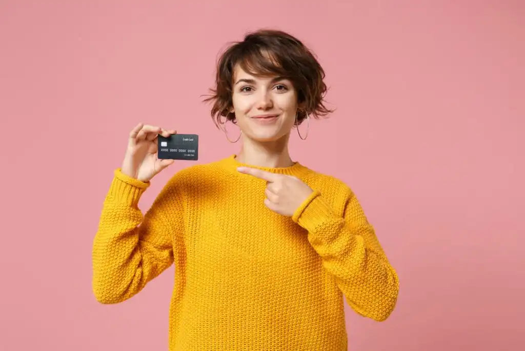 A smiling person in a yellow sweater stands against a pink background, holding a black credit card in one hand and pointing to it with the other.