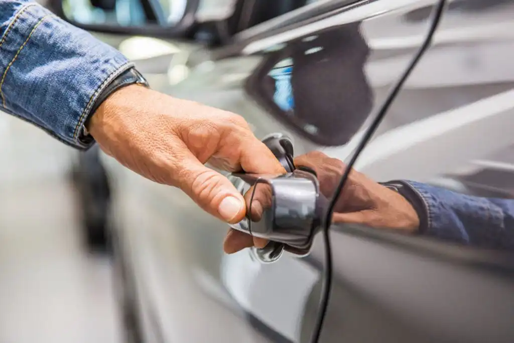 A close-up of a person’s hand, wearing a denim jacket, opening the door of a silver car. The focus is on the hand gripping the car door handle.