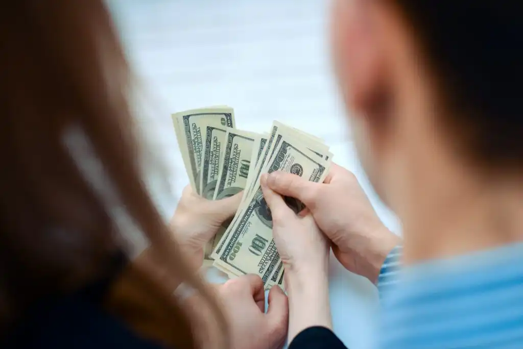 Two people, seen from behind, hold and count multiple $100 bills together, with their hands overlapping slightly, suggesting a financial transaction or exchange of money.