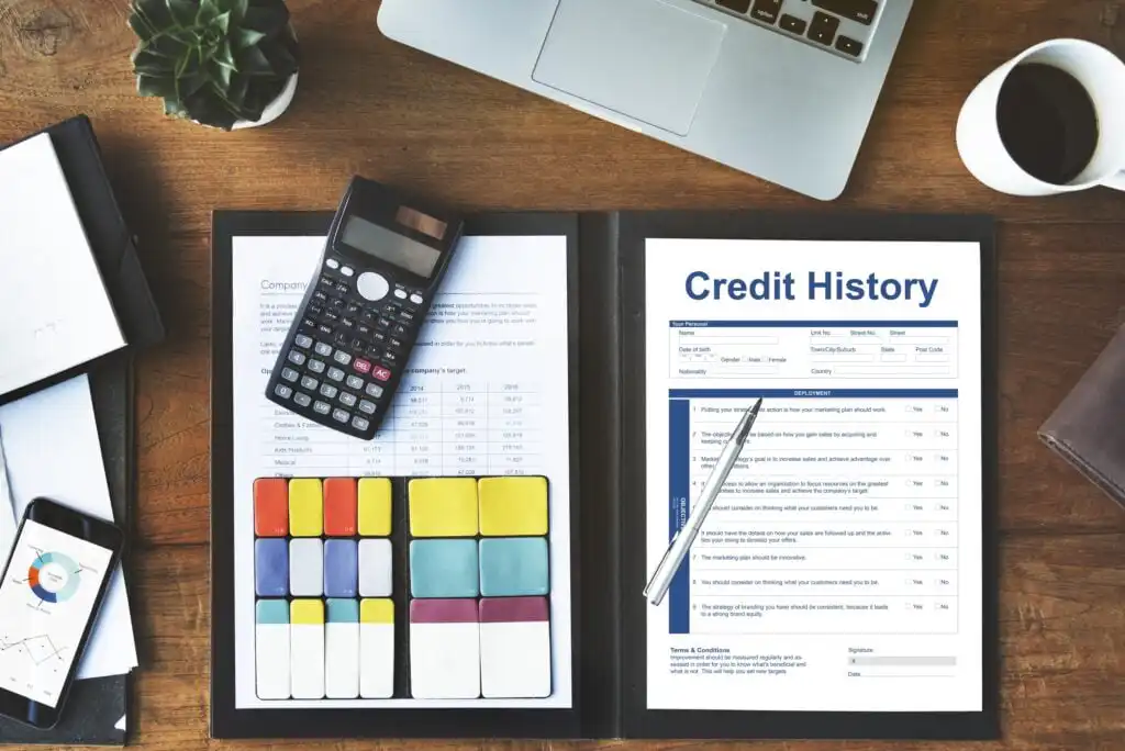 An open folder on a wooden desk displays a credit history report and colorful sticky notes. Nearby are a calculator, smartphone, laptop, a plant, and a cup of coffee. A pen rests on the report.