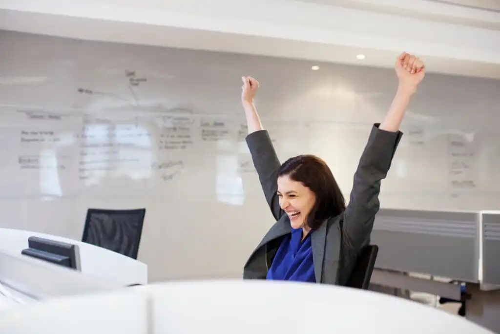 A woman in business attire sits at a desk in an office, smiling broadly and raising both arms in triumph. She appears happy and excited, with a whiteboard filled with notes in the background.