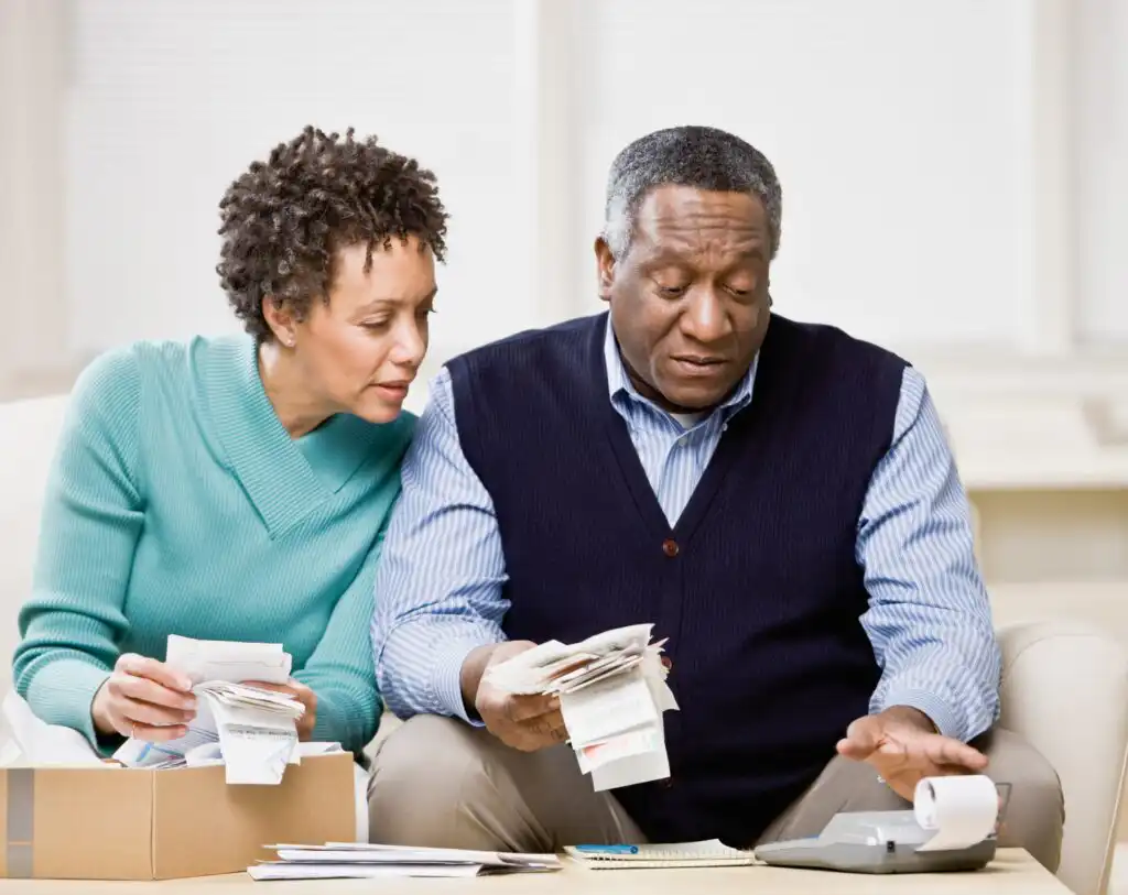 An older couple sits on a couch reviewing bills and receipts together, with a box of papers on the table and the man using a calculator. They appear focused and concerned about their finances.