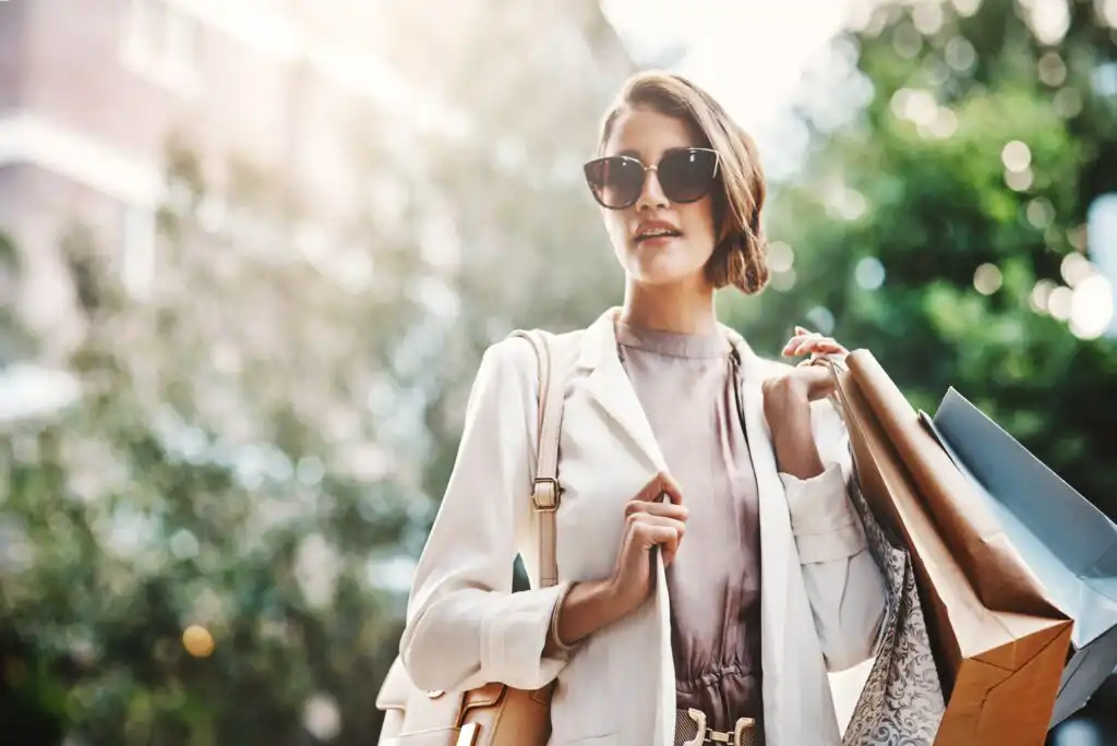 A stylish woman wearing sunglasses and a white blazer holds several shopping bags while walking outdoors on a sunny day, with greenery and sunlight in the background.