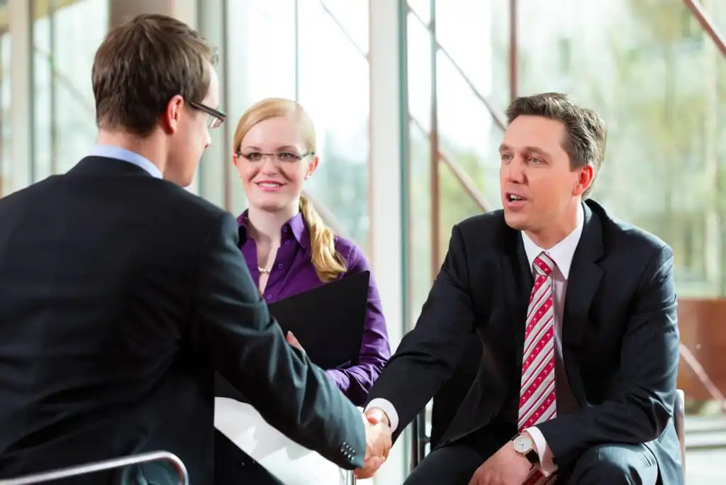 Three professionals in business attire are in a meeting. Two men are shaking hands while a woman sits between them, smiling and holding a black folder. Large windows and natural light are visible in the background.