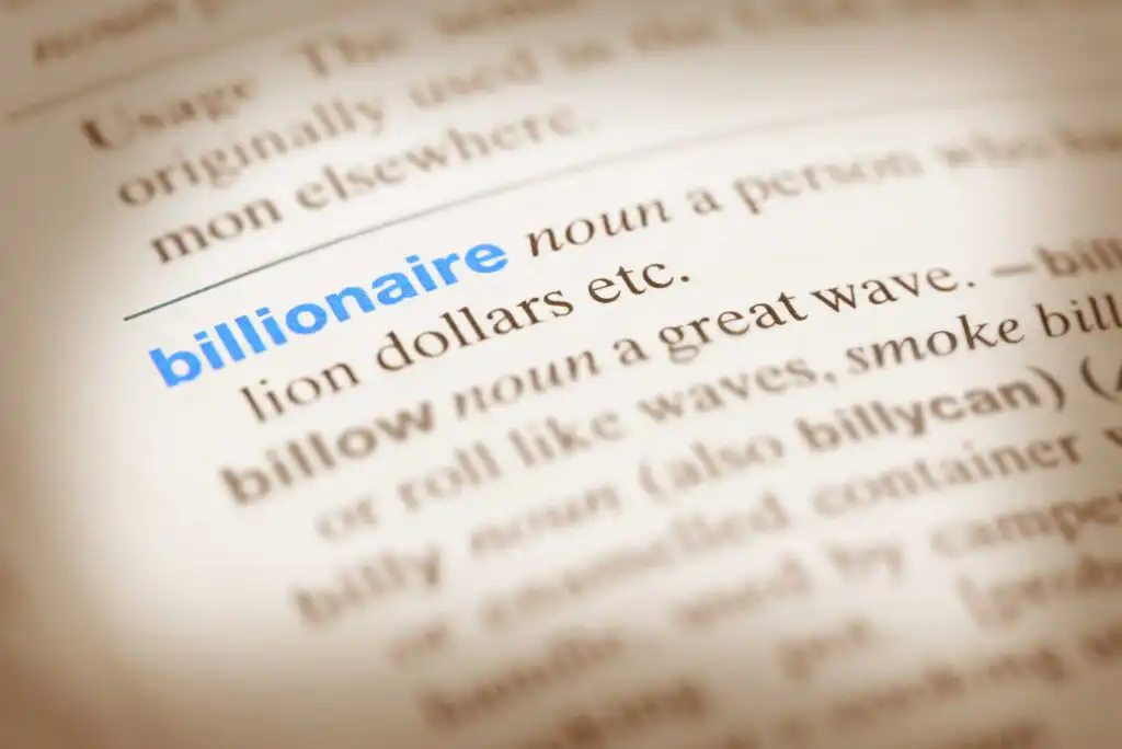 A close-up of a dictionary page highlighting the word billionaire, shown in bold blue text, with a partial definition visible next to it. The surrounding text is slightly blurred.