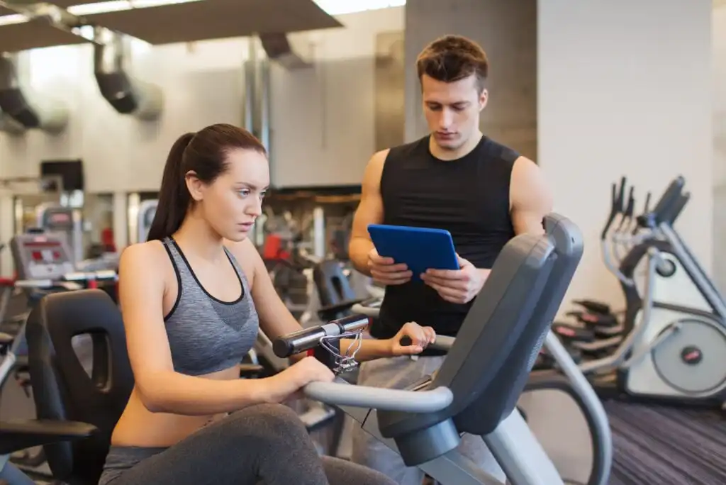 A woman using a stationary bike in a gym while a man in athletic wear stands beside her, holding a tablet and observing her progress. Exercise equipment is visible in the background.
