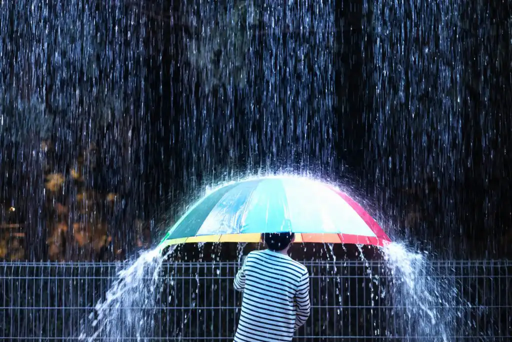 A person wearing a striped shirt stands under a large, colorful umbrella, sheltering from heavy rain. Water streams down the umbrella’s sides, creating a curtain effect. The scene is lit dramatically against a dark background.
