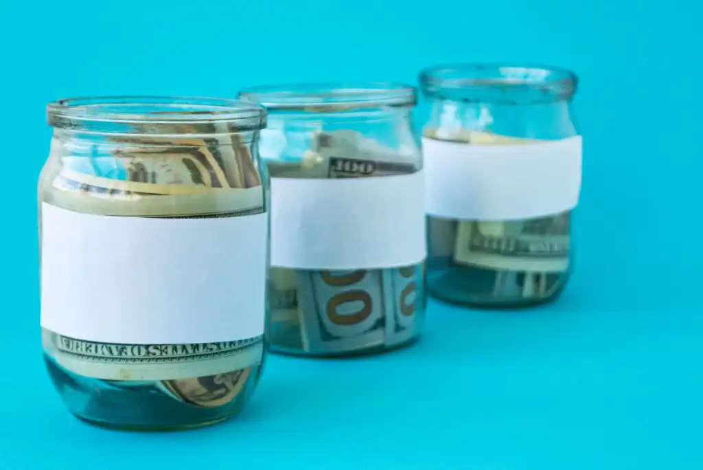 Three glass jars with blank white labels, each filled with U.S. dollar bills, are arranged in a row against a bright blue background.