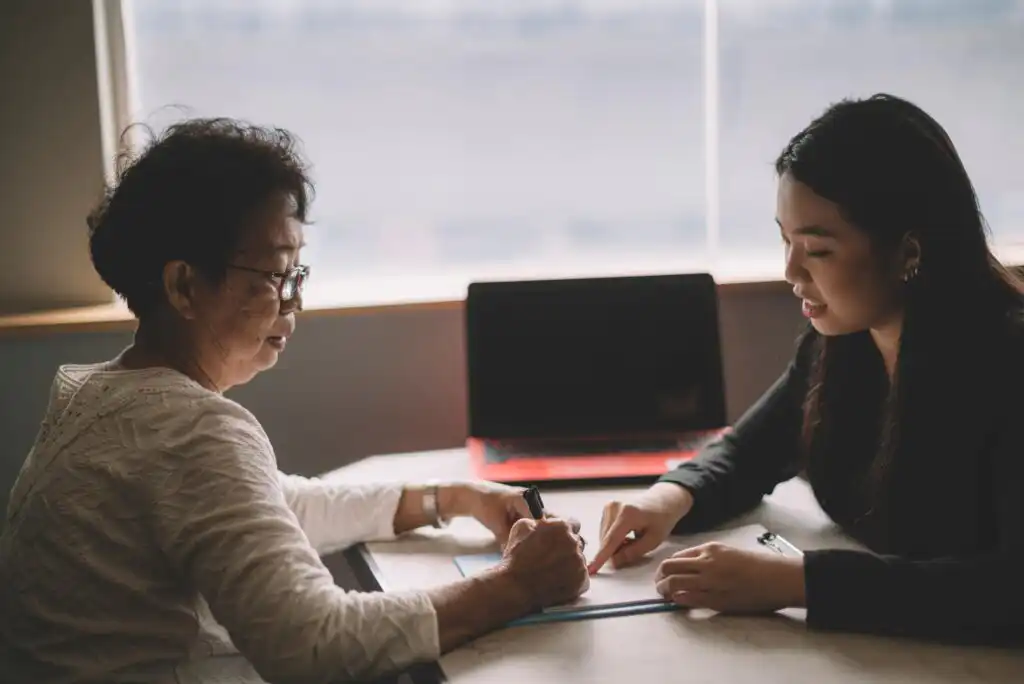 An older woman and a younger woman sit at a table discussing documents. The older woman holds a pen, while the younger woman points to the paperwork. A laptop sits closed in the background.