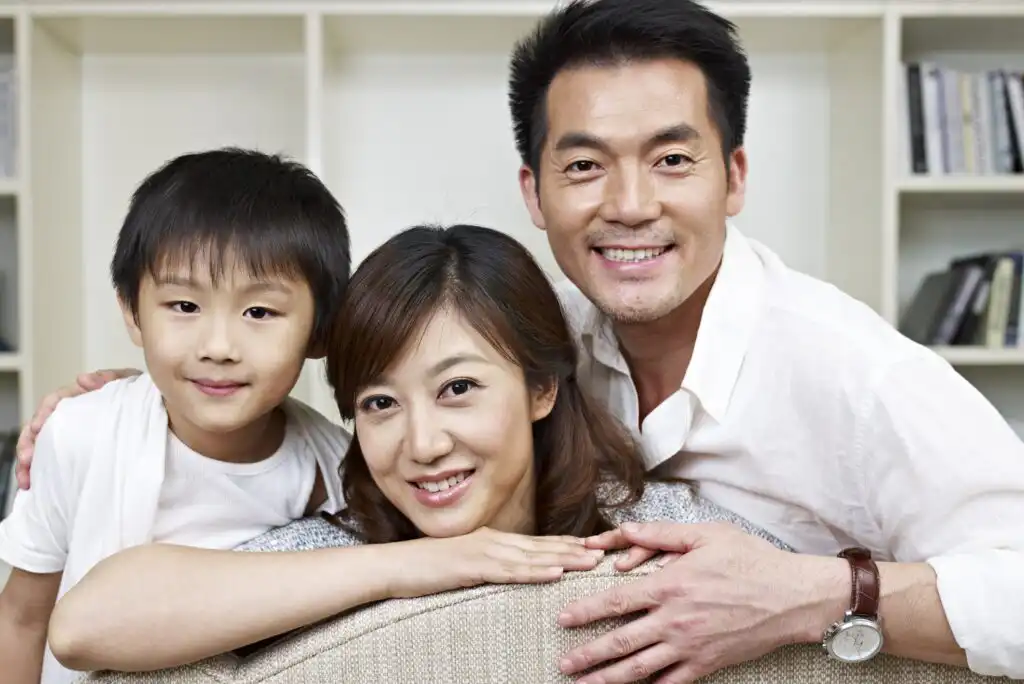 A smiling family of three, including a man, woman, and young boy, pose together indoors in front of a bookshelf. The adults are wearing white shirts, and the boy is leaning on the back of a couch.