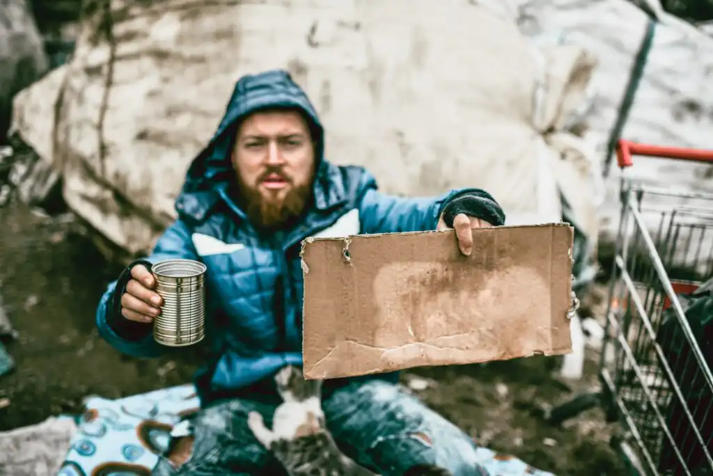 A man in a blue jacket and gloves sits outdoors, holding a blank cardboard sign in one hand and an empty tin can in the other, suggesting he is asking for help or donations.