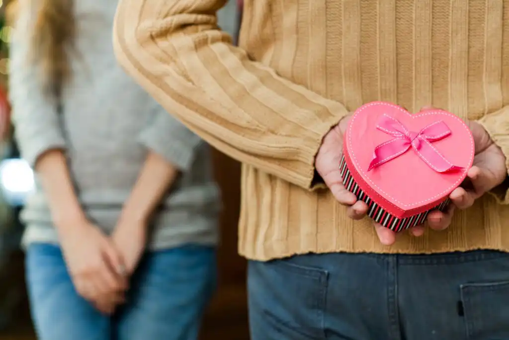 A person in a brown sweater holds a heart-shaped gift box with a pink bow behind their back, while another person stands in the background with hands clasped.