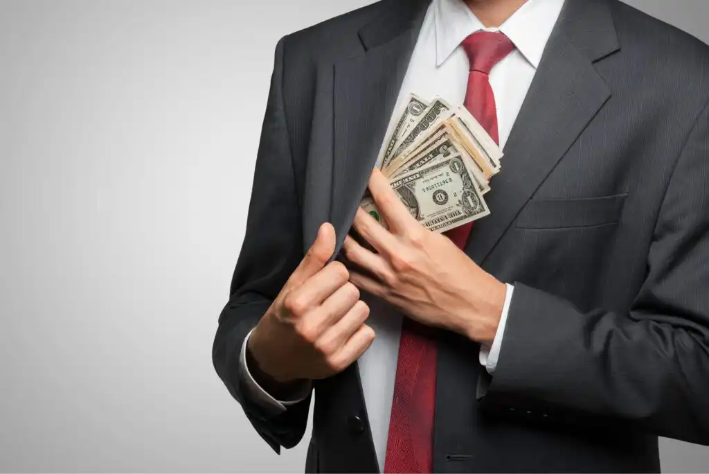 A man in a suit and red tie puts several U.S. dollar bills into his jacket pocket, partially hiding the money. The image focuses on his torso and hands, not showing his face.