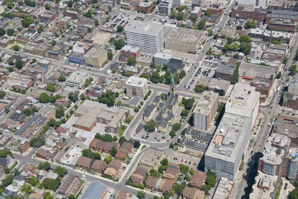 Aerial view of a city neighborhood with a mix of residential and commercial buildings, tree-lined streets, parking lots, and green spaces on a sunny day.
