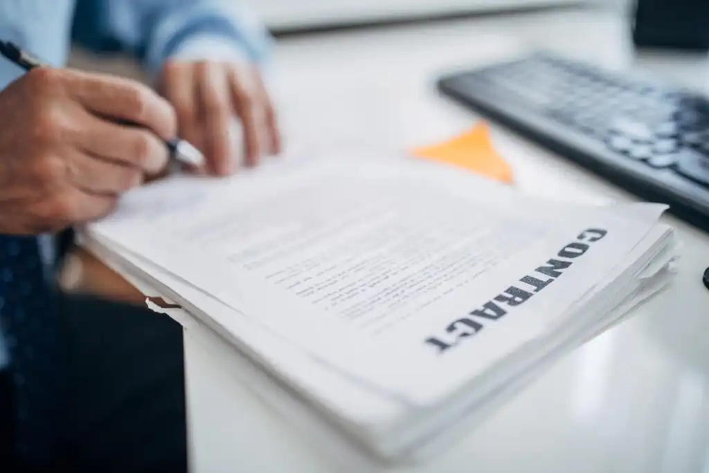 A person signs a paper document labeled CONTRACT at a desk, with a keyboard and some papers visible in the background.
