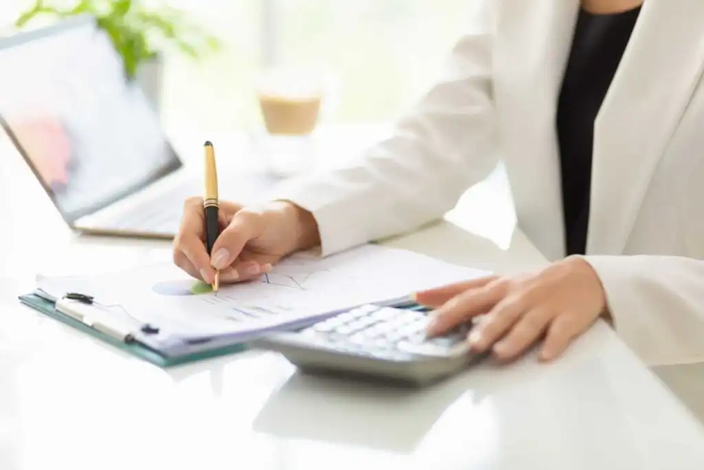 A person in a white blazer is using a calculator and writing on documents with charts at a desk. A laptop and a blurred coffee cup are in the background.