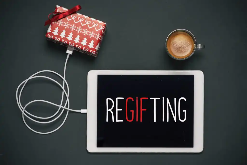 A tablet on a dark surface displays the word “REGIFTING,” with “GIFT” in red. Next to it are a wrapped present, a cup of coffee, and white earphones connected to the tablet.