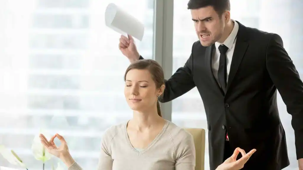 A calm woman practices meditation at her desk while a man in a suit angrily yells at her, holding papers in his hand. They are in an office setting with large windows in the background.
