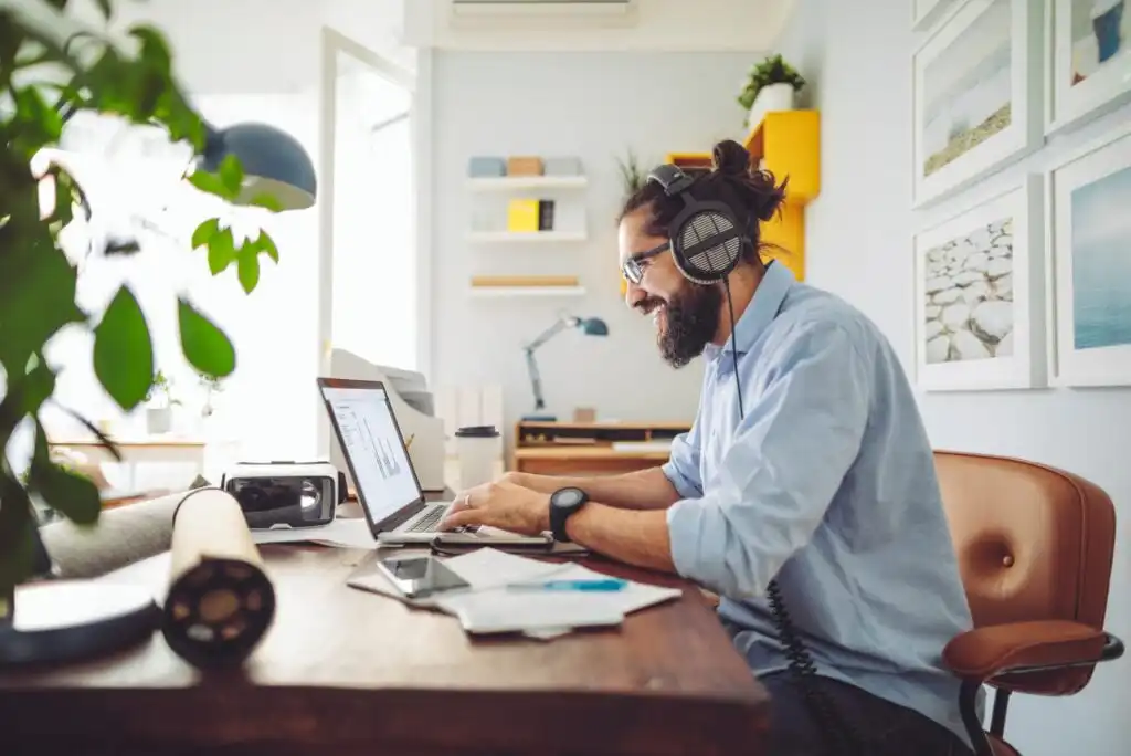 A man with headphones and glasses works on a laptop at a desk in a bright, modern home office, surrounded by papers, plants, and shelves with books and decor.