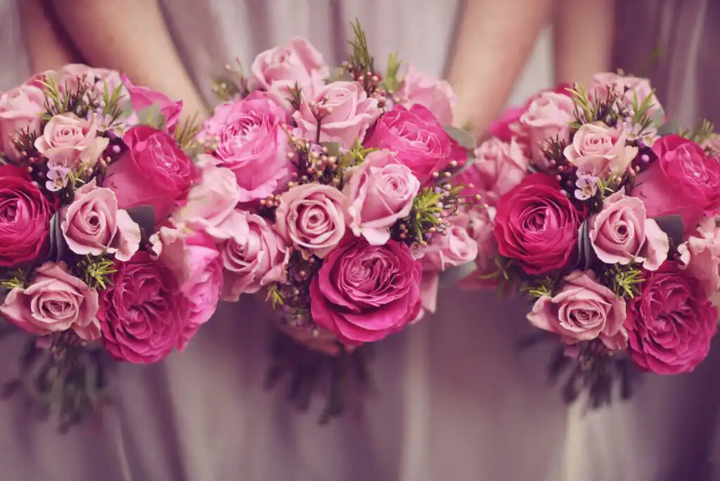Three bouquets of pink and magenta roses with greenery are held by people in light-colored dresses, creating a soft and romantic floral arrangement.