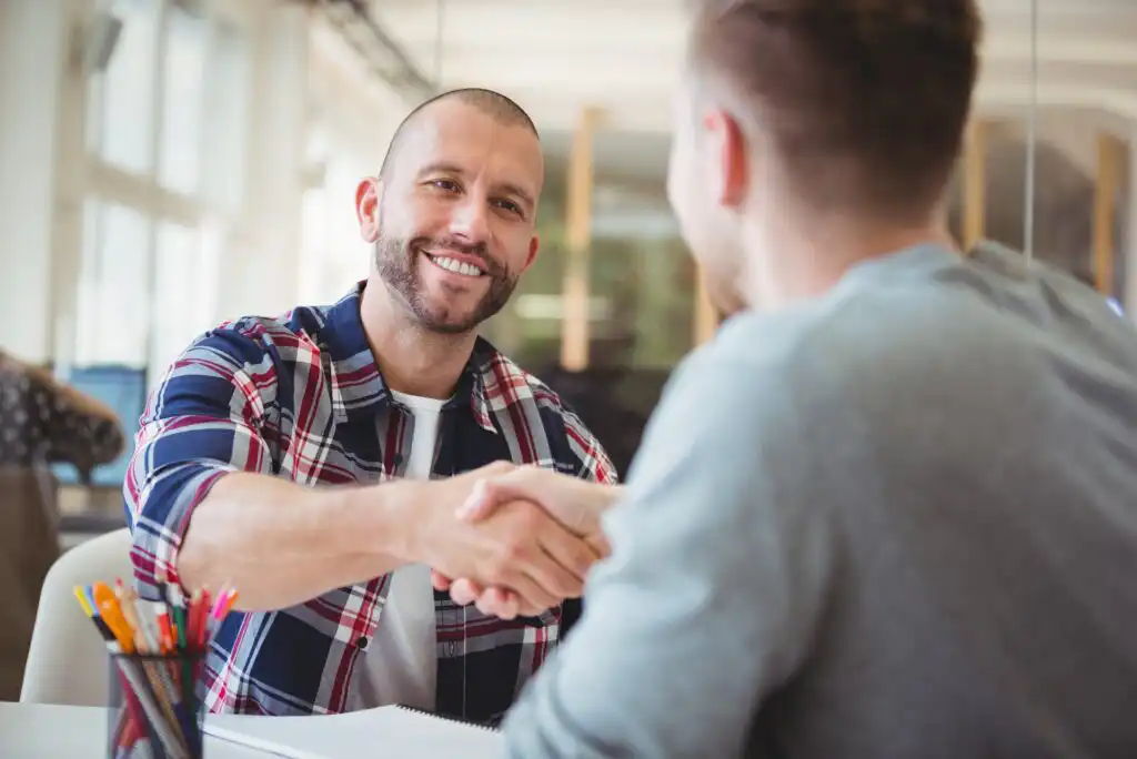 Two men are sitting at a desk in an office, smiling and shaking hands. One man is wearing a plaid shirt and the other is in a gray shirt. A cup filled with colorful pens is visible on the table.